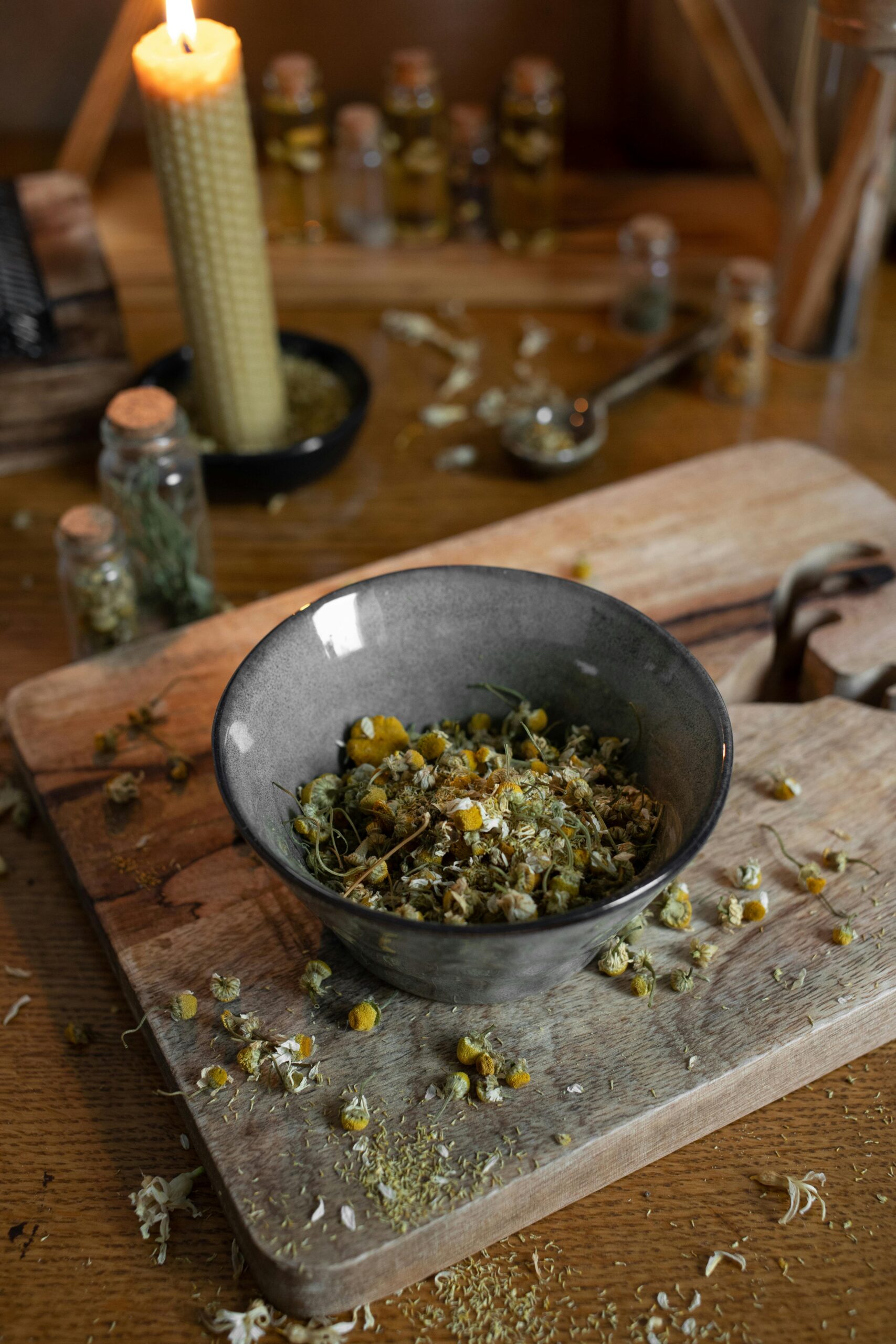 Dried chamomile flowers in a bowl on a wooden board, surrounded by herb jars and a beeswax candle